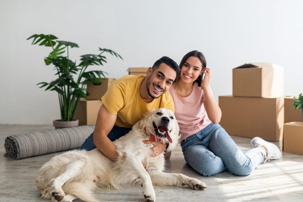 Man, woman, and dog smiling in front of moving boxes in apartment room.