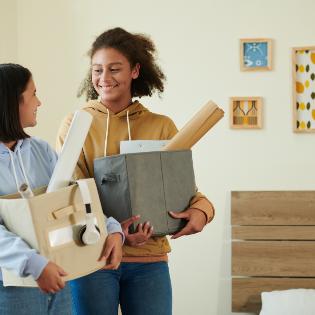 Two roommates carrying boxes into their apartment while smiling at each other.