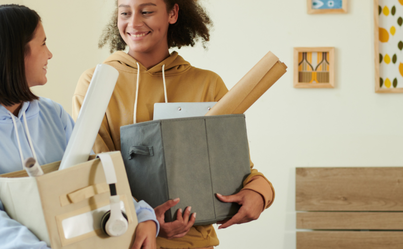 Two roommates carrying boxes into their apartment while smiling at each other.